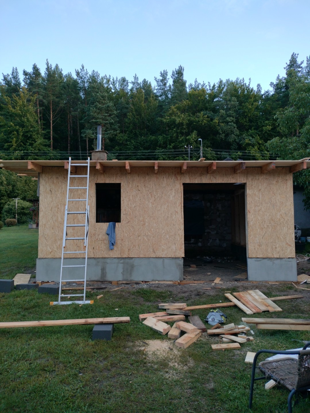 Budowa parterowego domu z drewnianą konstrukcją szkieletową, widoczna drabina oparta o ścianę, resztki materiałów budowlanych na trawie i las w tle.