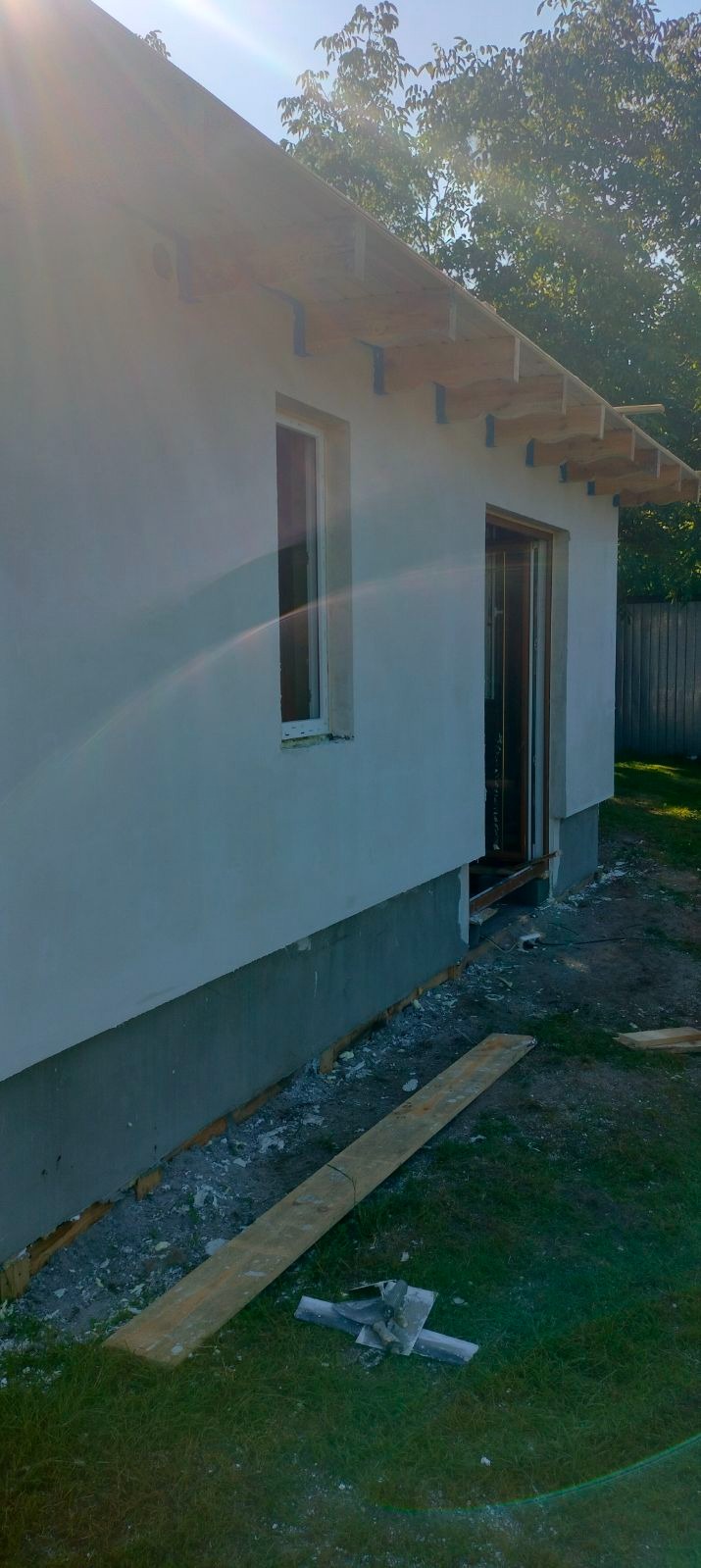Ściana domu z oknem i otwartymi drzwiami, w trakcie prac wykończeniowych, widoczne drewniane elementy konstrukcji dachu, fragment fundamentu i deska na trawie.
