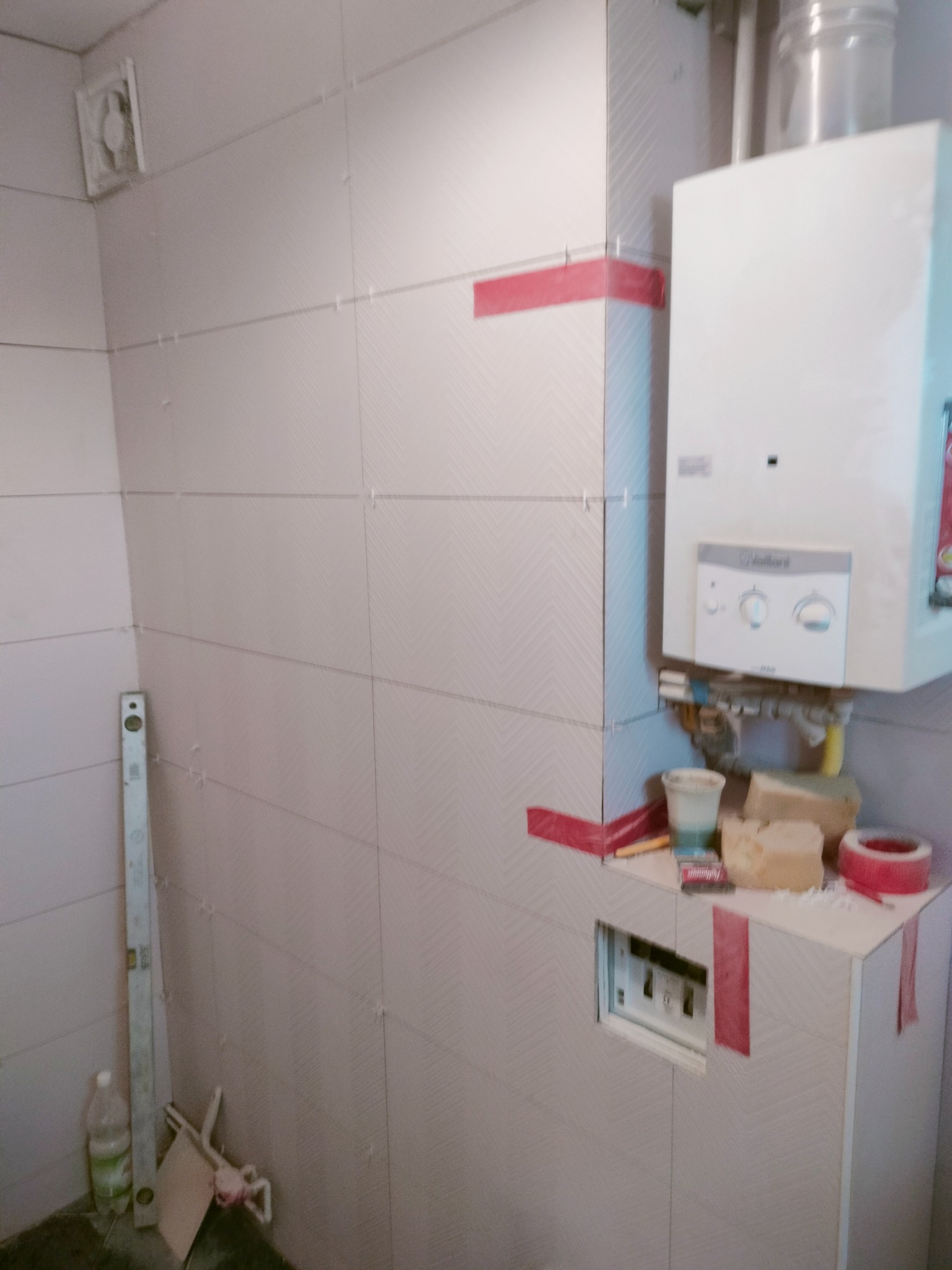 Łazienka w trakcie remontu, z częściowo położonymi jasnymi płytkami chevron, poziomica oparta o ścianę, widoczny piec gazowy i przygotowane materiały budowlane.