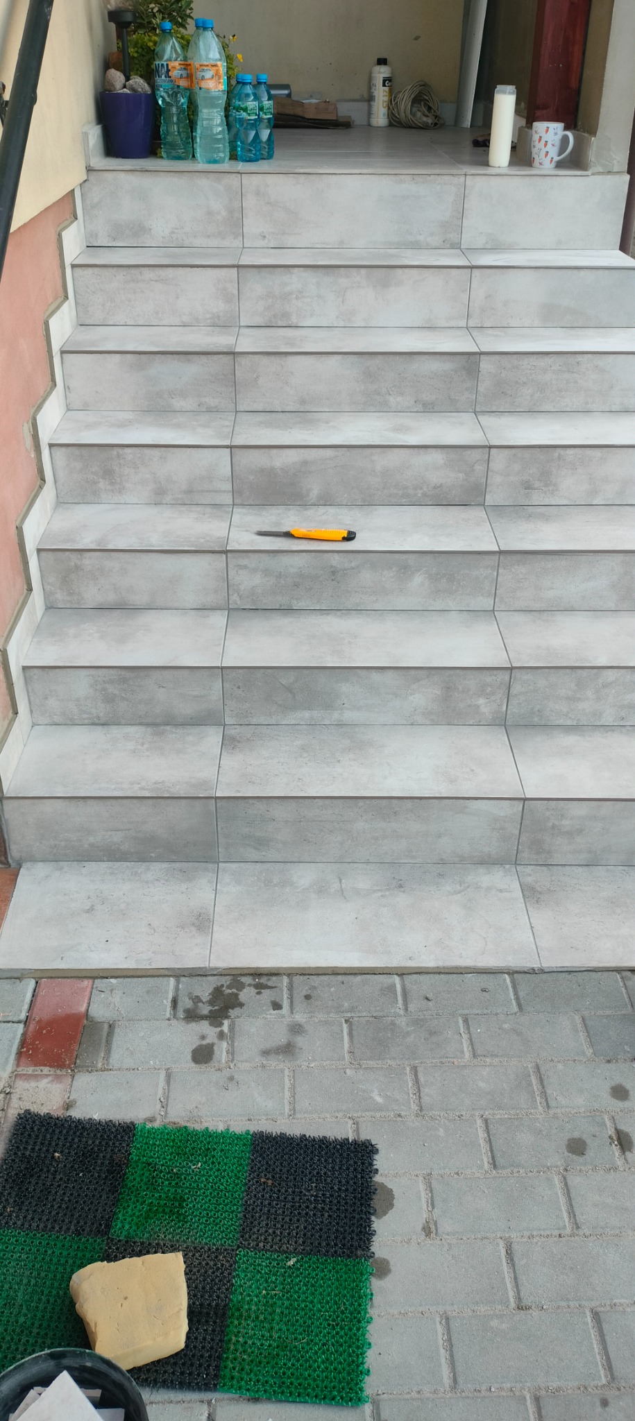 Wykończone schody zewnętrzne z szarych płytek, na jednym ze stopni leży nóż monterski z żółtą rączką. U góry w tle widać butelki z wodą i inne akcesoria budowlane, a u dołu wycieraczkę i resztki...