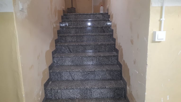 Schody wyłożone szarym, nakrapianym granitem, z widocznymi śladami świeżo położonych gładzi na ścianach w klatce schodowej.