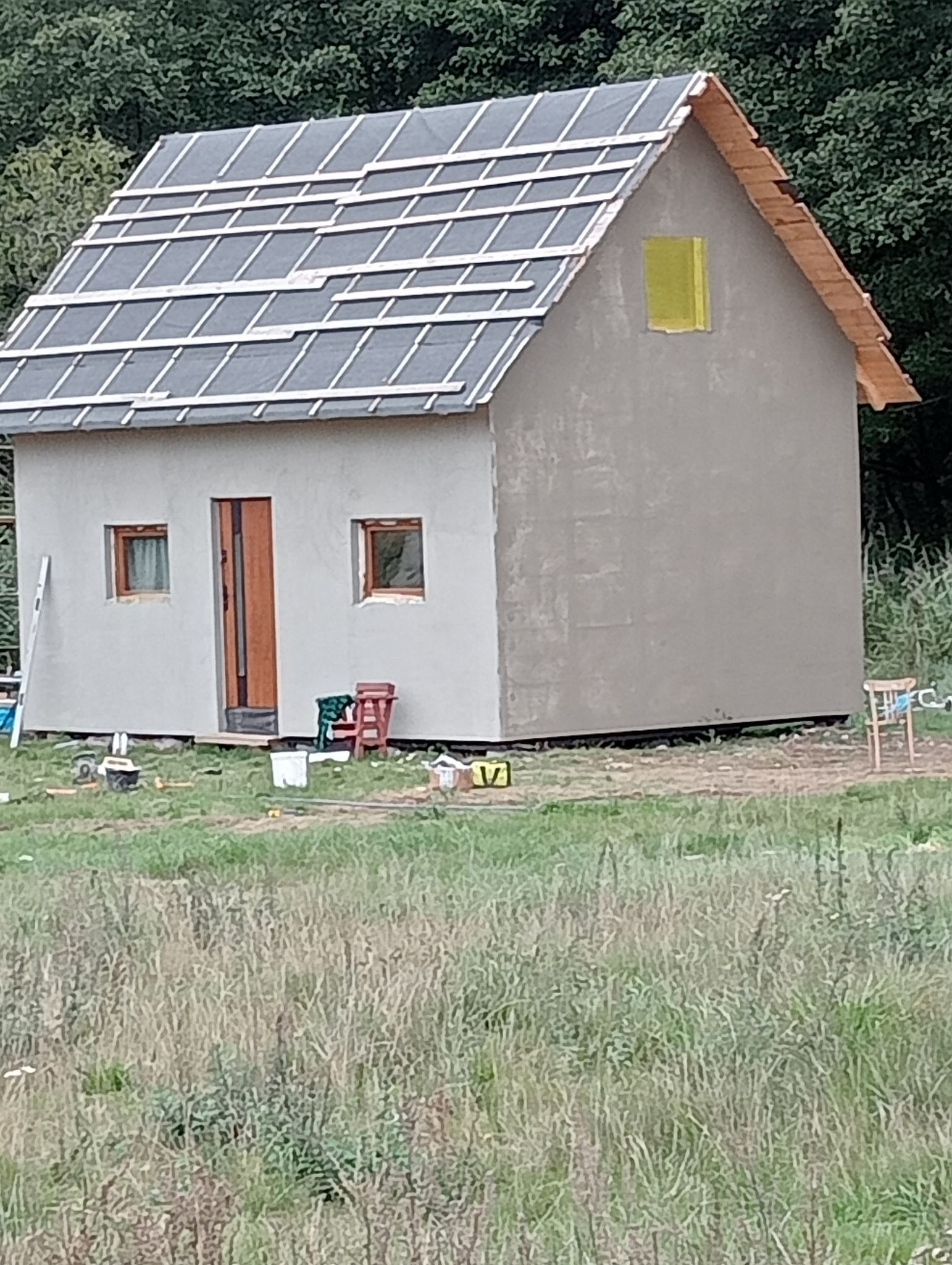 Niewielki, szary dom w stanie surowym otwartym, z częściowo położonym dachem z papy i listew kontrłat, z zamurowanym oknem zabezpieczonym folią, na tle zieleni.
