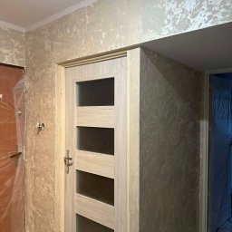 VIRA-BUD SVITLANA MALYSHENKO - Wykończone ściany z efektem dekoracyjnym w odcieniach beżu i szarości, widoczna drewniana ościeżnica z przeszklonymi panelami i zabezpieczone folią drzwi po lewej.