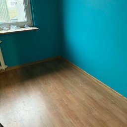 VIRA-BUD SVITLANA MALYSHENKO - Turkusowa ściana i panele podłogowe w pokoju po remoncie. Widoczne okno z parapetem i grzejnik. Świeżo pomalowane ściany i ułożona podłoga.