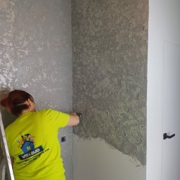 VIRA-BUD SVITLANA MALYSHENKO - Pracownik w neonowej koszulce nanosi szpachelką dekoracyjną, szarą masę na ścianę w rogu pokoju. Widoczna drabina i białe drzwi.