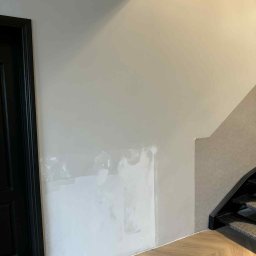 VIRA-BUD SVITLANA MALYSHENKO - Ściana w trakcie przygotowania do malowania, widoczne ślady szpachlowania. Parkiet ułożony w jodełkę, czarne drzwi po lewej stronie kadru, fragment schodów po prawej.