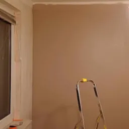 Pokój po szpachlowaniu ścian na kolor beżowy, widoczna drabina aluminiowa z żółtymi elementami, okno z zasłoniętą roletą, na parapecie narzędzia i taśma malarska.