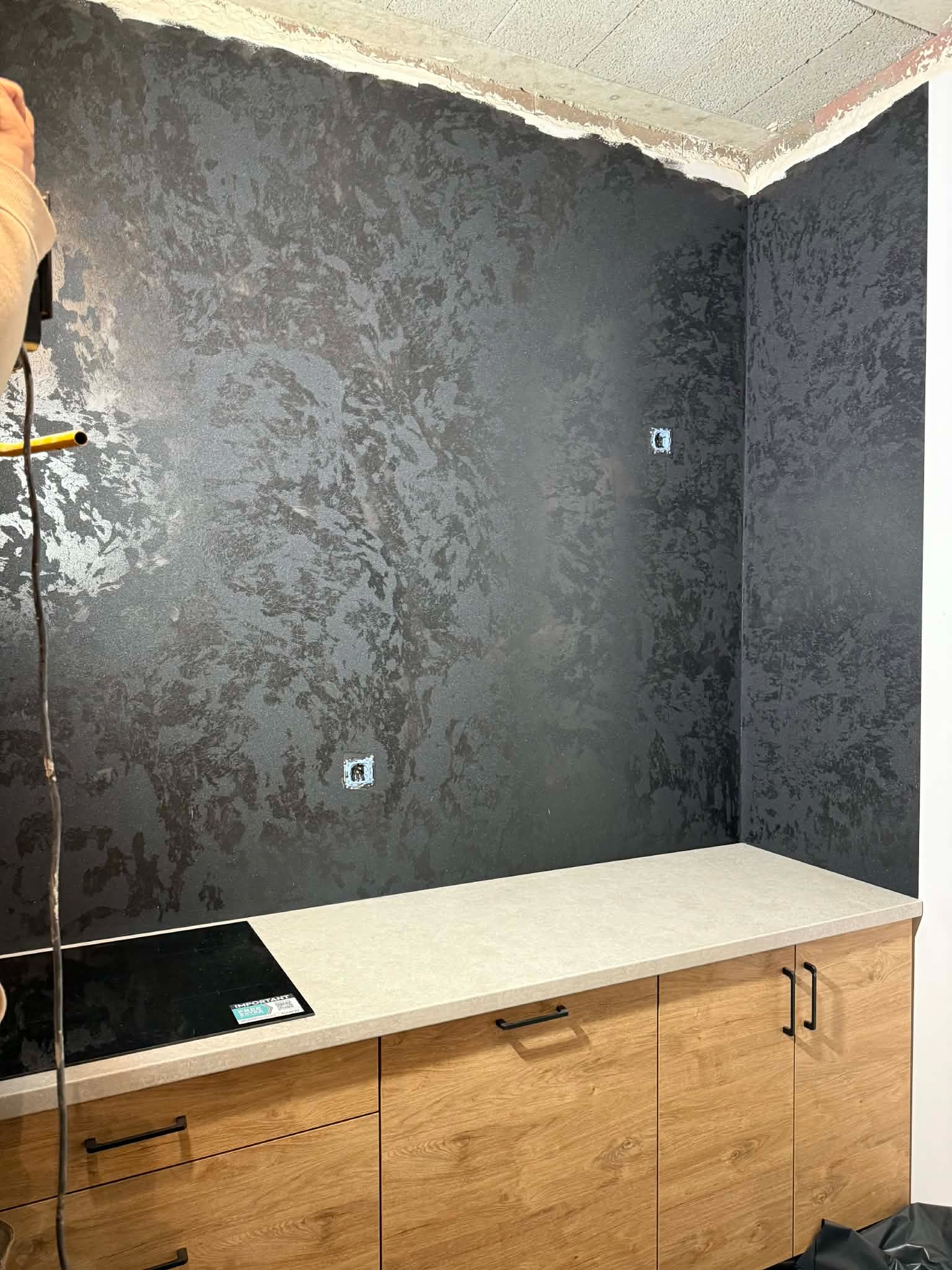 Ciemna ściana z efektem dekoracyjnym w kuchni, widoczne gniazdka elektryczne i fragment zabudowy kuchennej z drewnianymi frontami. Wykończenie wnętrza w nowoczesnym stylu.