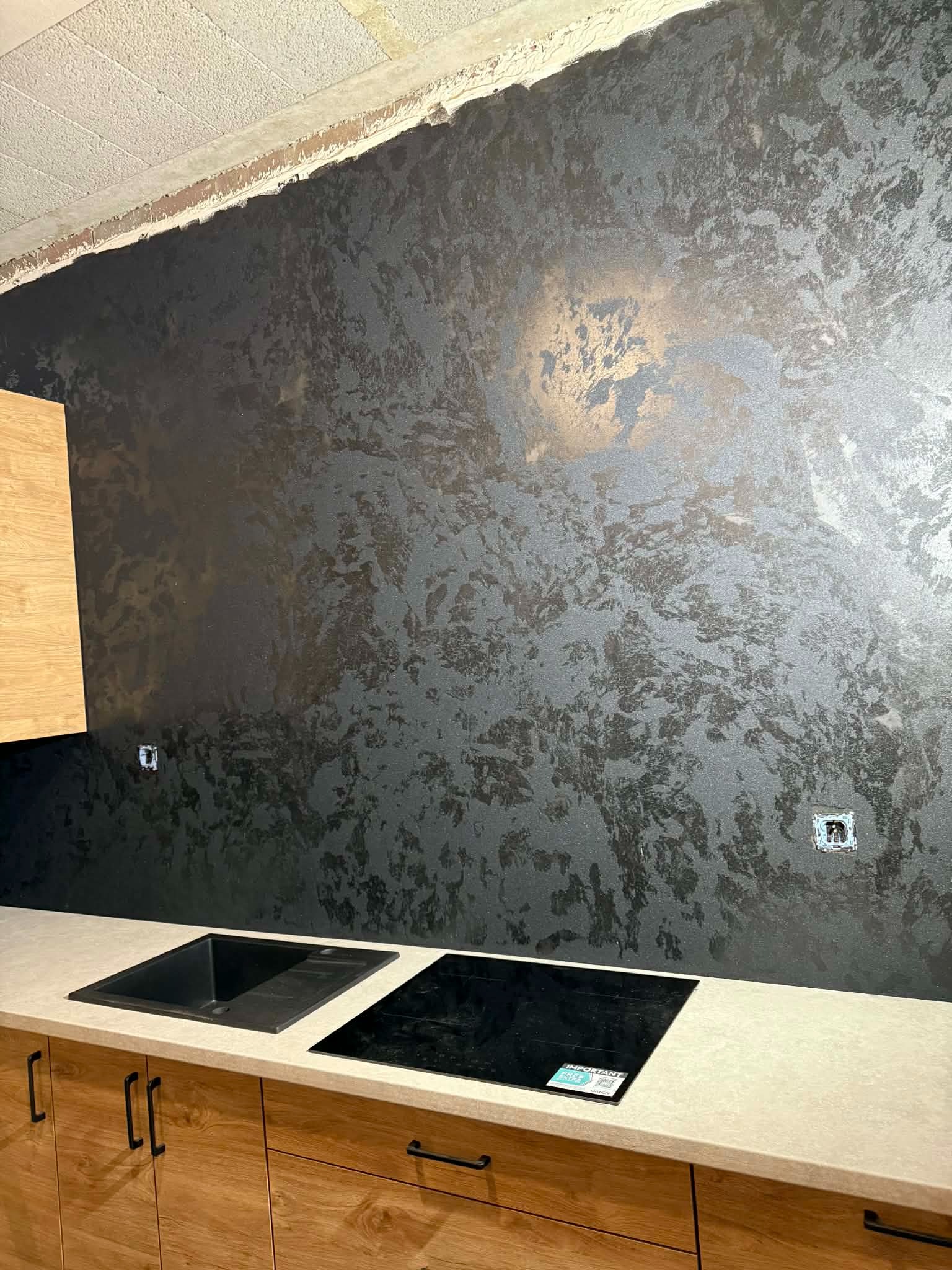 Ściana w kuchni z dekoracyjną, ciemną fakturą, widoczne gniazdka elektryczne. Część zabudowy kuchennej z drewnianymi frontami i blatem.