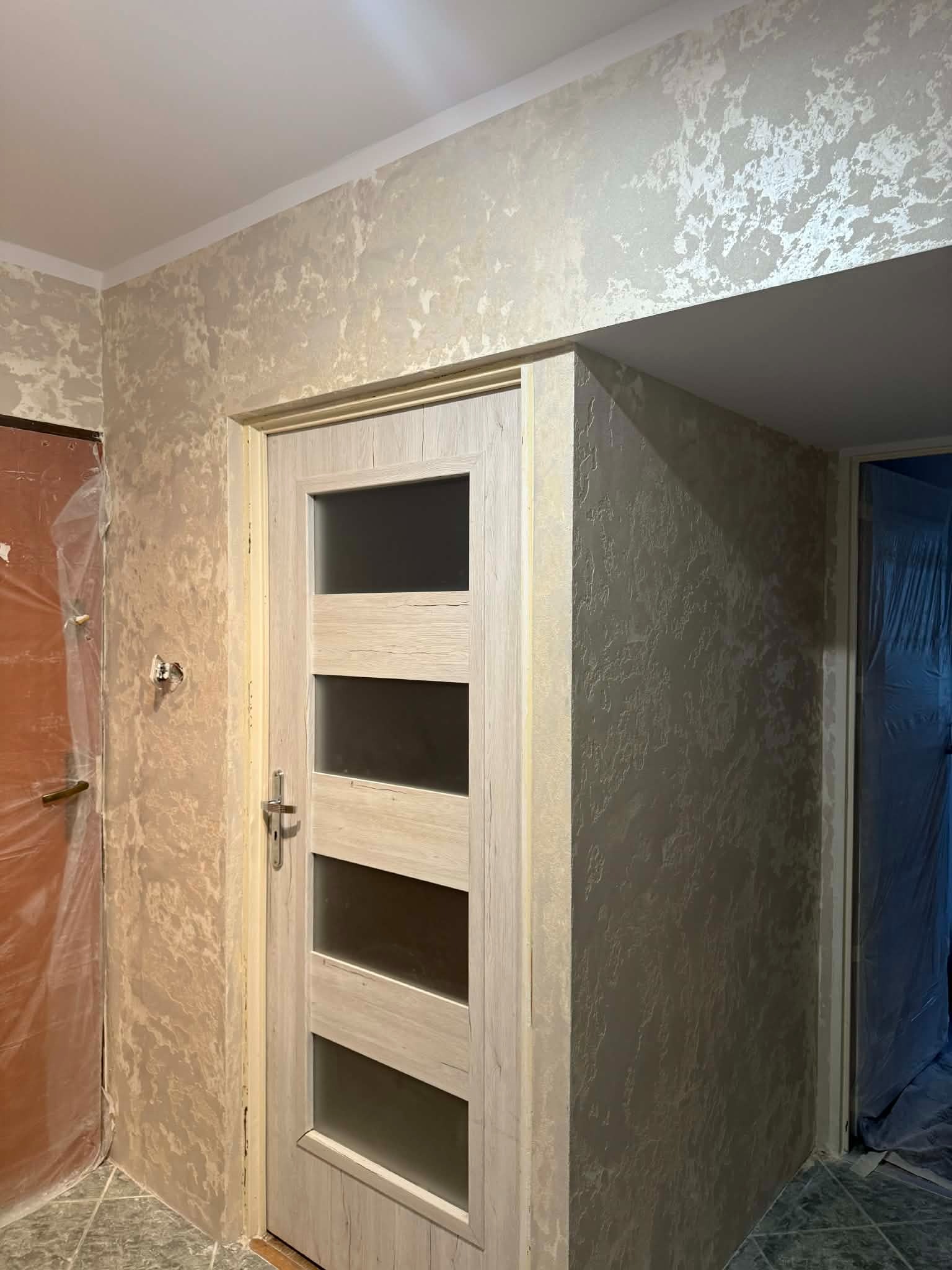 Wykończone ściany z efektem dekoracyjnym w odcieniach beżu i szarości, widoczna drewniana ościeżnica z przeszklonymi panelami i zabezpieczone folią drzwi po lewej.