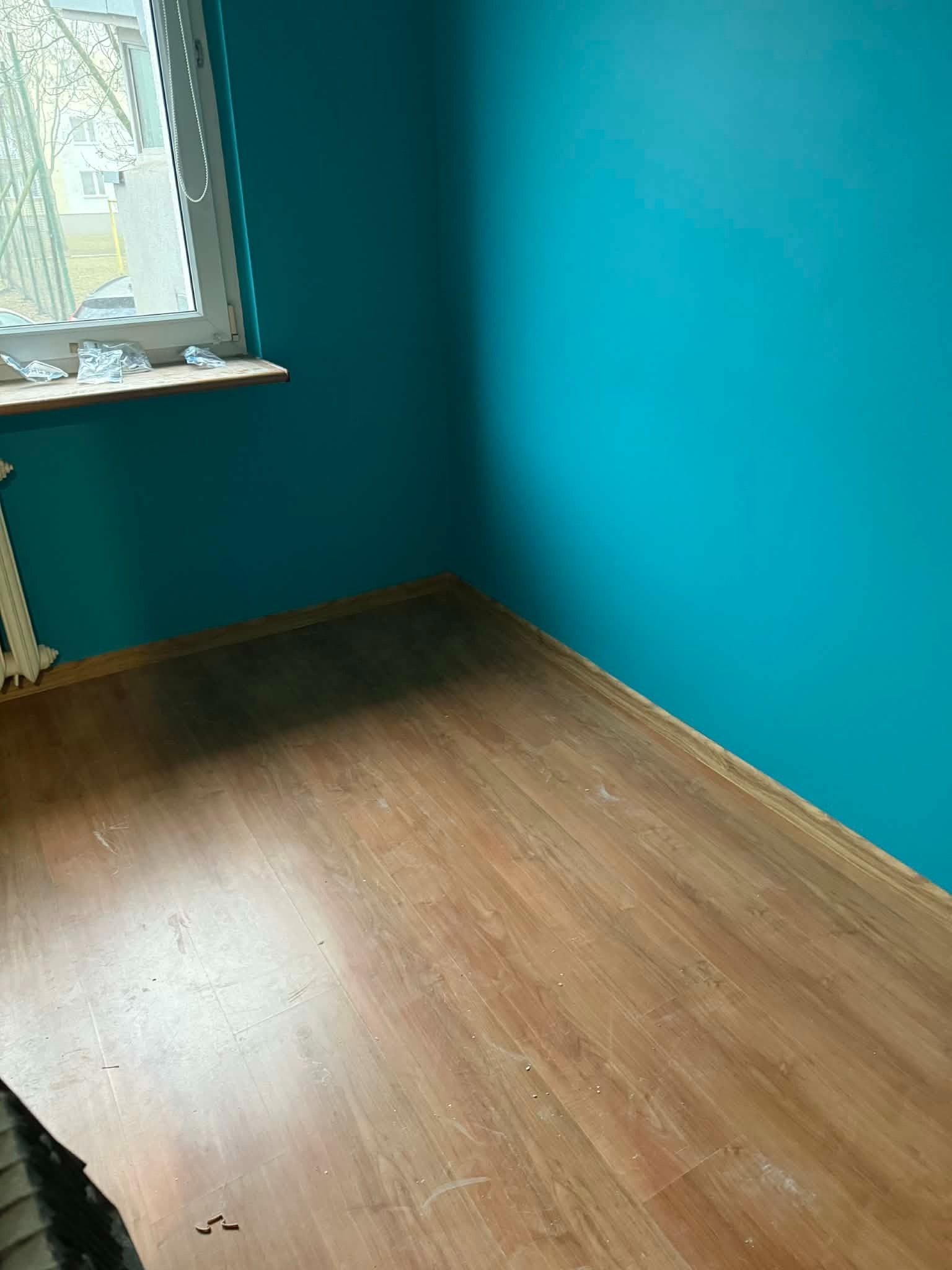 Turkusowa ściana i panele podłogowe w pokoju po remoncie. Widoczne okno z parapetem i grzejnik. Świeżo pomalowane ściany i ułożona podłoga.