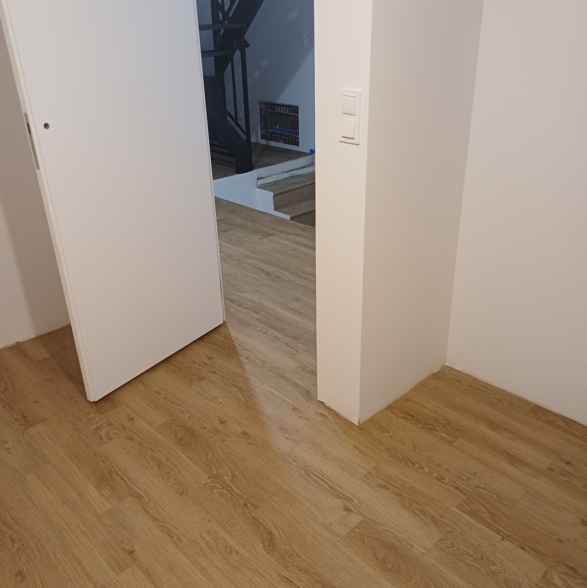 Jasne panele podłogowe w korytarzu, widok na schody i otwarte drzwi. Świeżo pomalowane ściany w odcieniach bieli, nowoczesny włącznik światła.