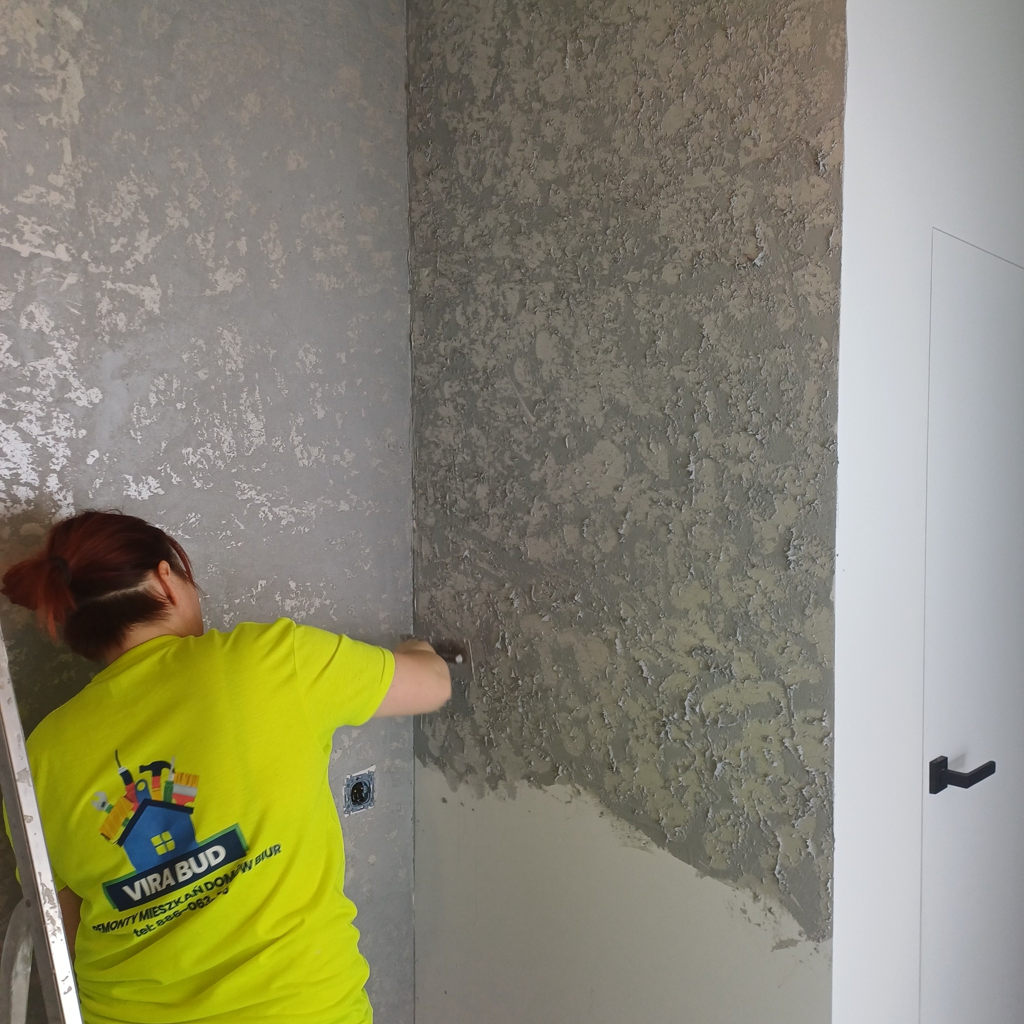 Pracownik w neonowej koszulce nanosi szpachelką dekoracyjną, szarą masę na ścianę w rogu pokoju. Widoczna drabina i białe drzwi.