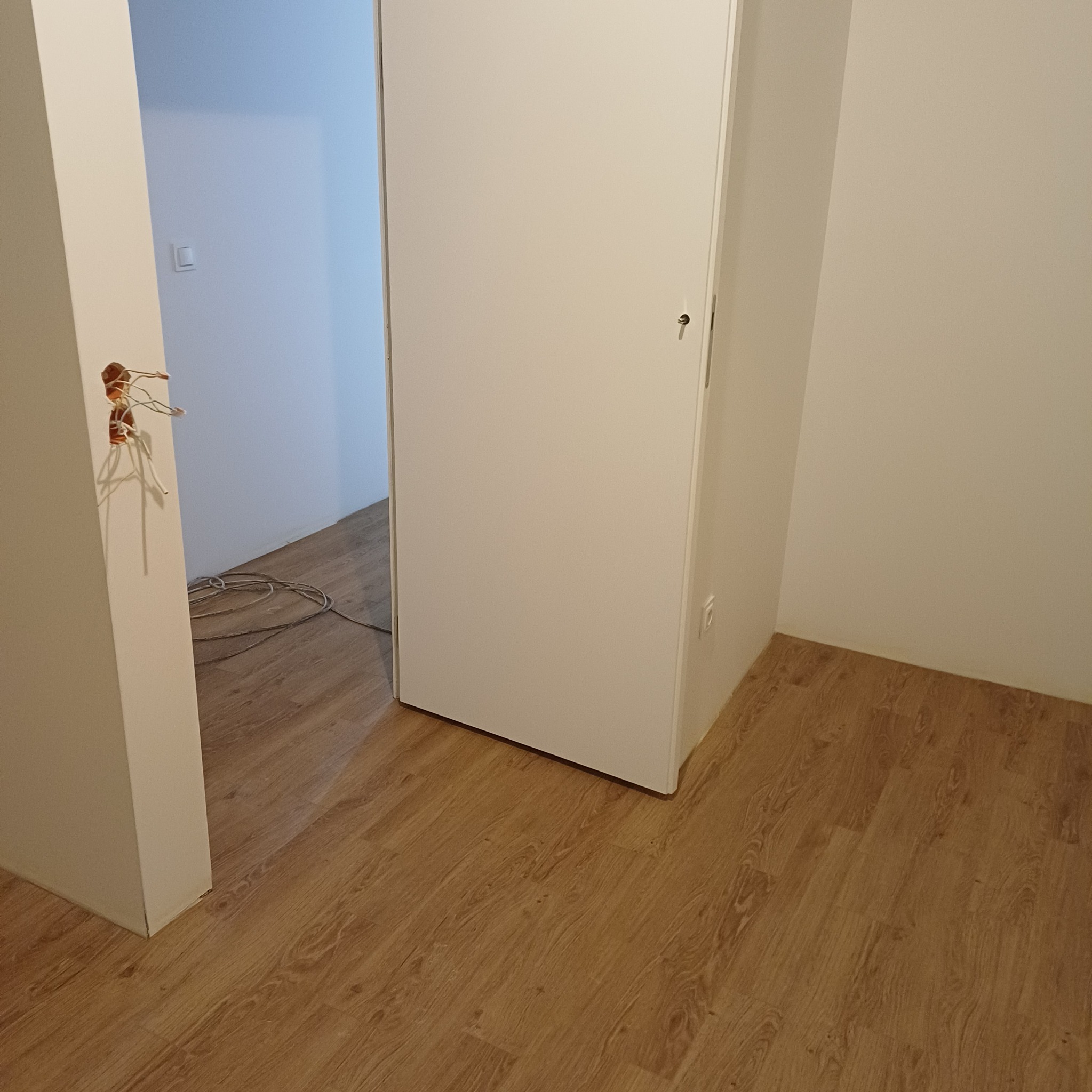 Wnętrze w trakcie remontu: widoczne kable elektryczne wystające ze ściany, świeżo położone panele podłogowe i białe ściany. Częściowo widoczne drzwi.