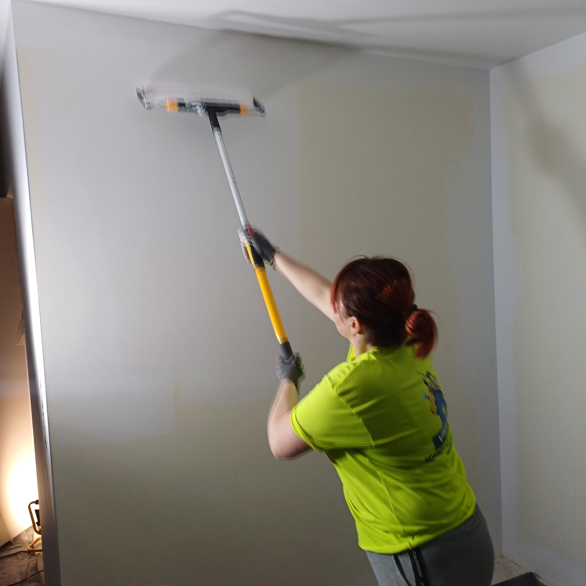 Kobieta w trakcie malowania ściany wałkiem na długim kiju. Widoczny fragment pomalowanej ściany i narożnik. Ubrana w roboczą koszulkę z logo firmy.