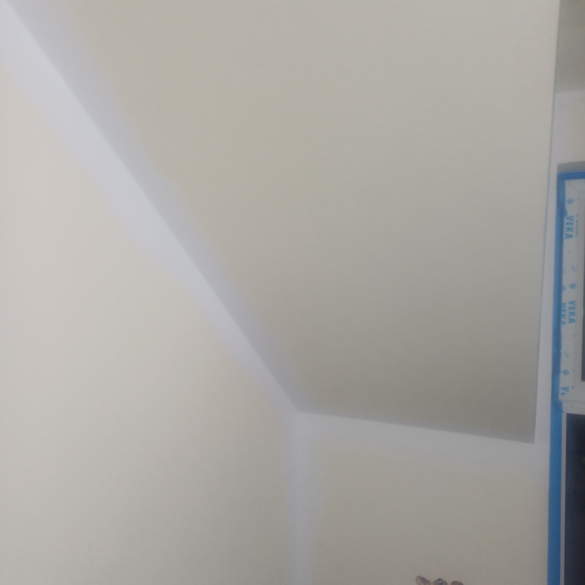 Ściana po gładzi i malowaniu w odcieniach bieli, widoczny fragment okna z niebieską ramą. Ujęcie z dołu, podkreślające gładką powierzchnię i kąt nachylenia ściany.