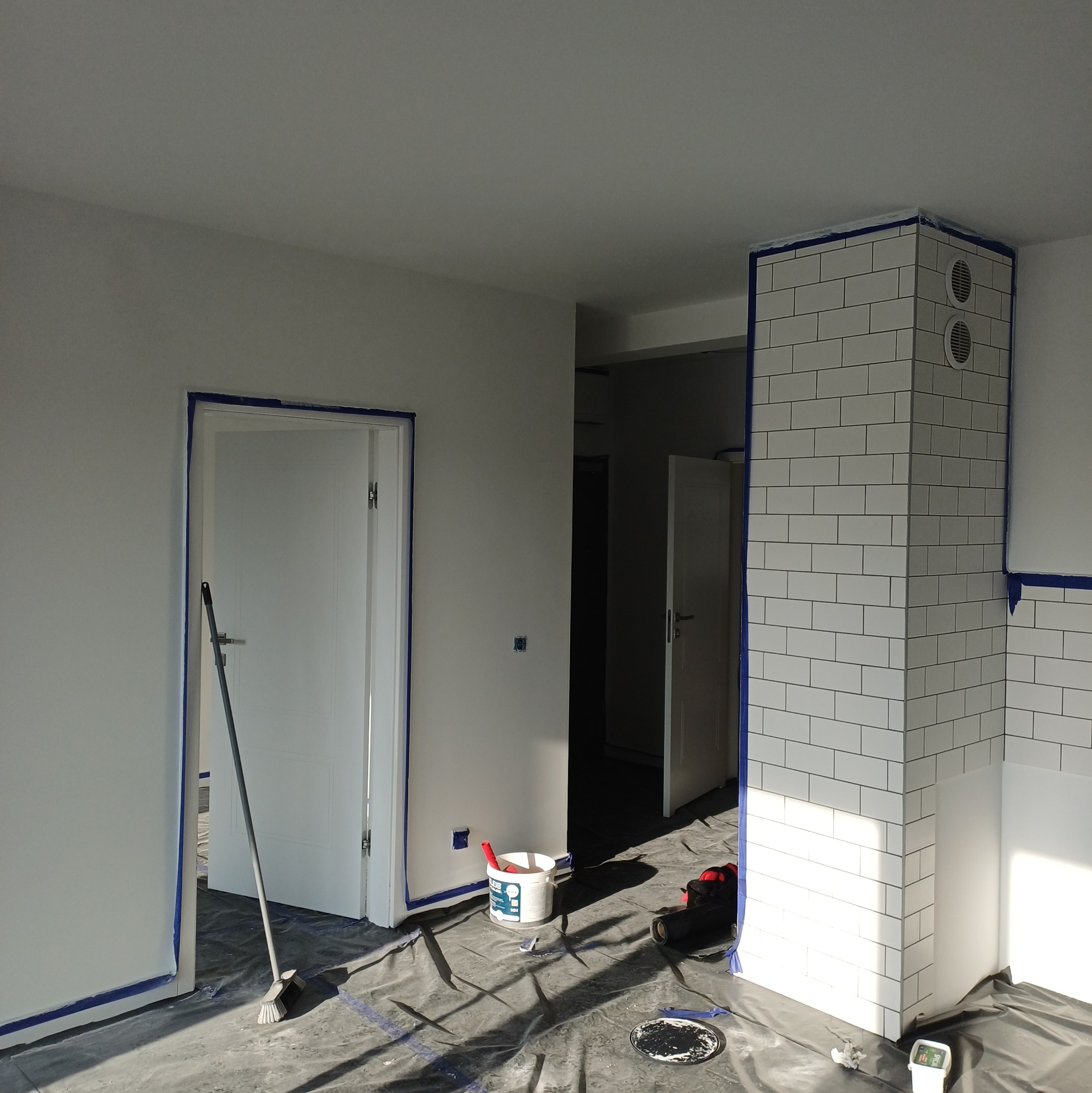 Przygotowania do malowania: zabezpieczone taśmą i folią drzwi, ściany i fragment z białymi płytkami. Widoczne narzędzia i wiadro z farbą. Jasne wnętrze w trakcie prac.