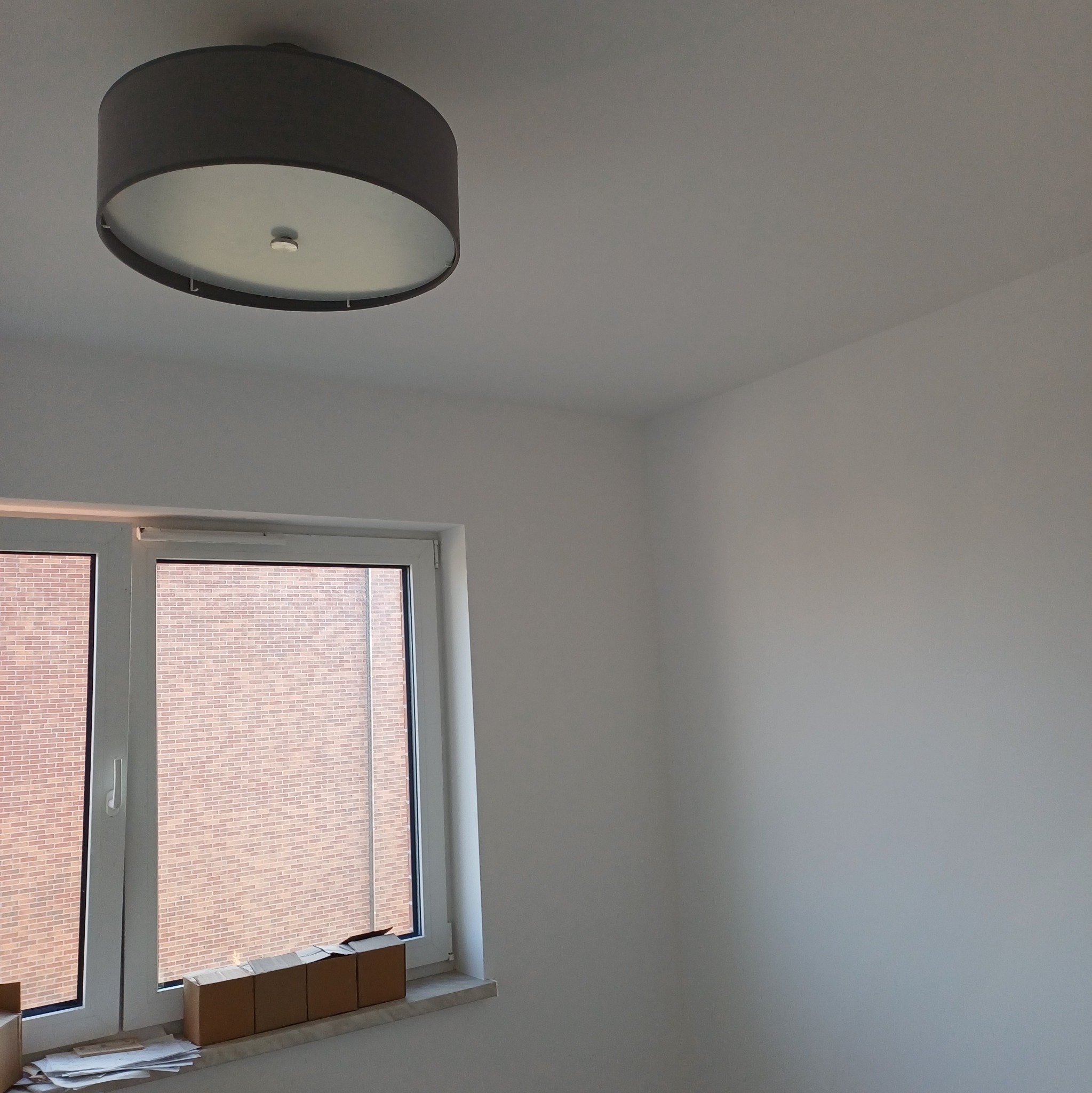 Świeżo pomalowane na biało ściany w pokoju w Gorzowie Wielkopolskim, widok na okno z parapetem, na którym leżą kartony i dokumenty. Na suficie okrągła lampa.