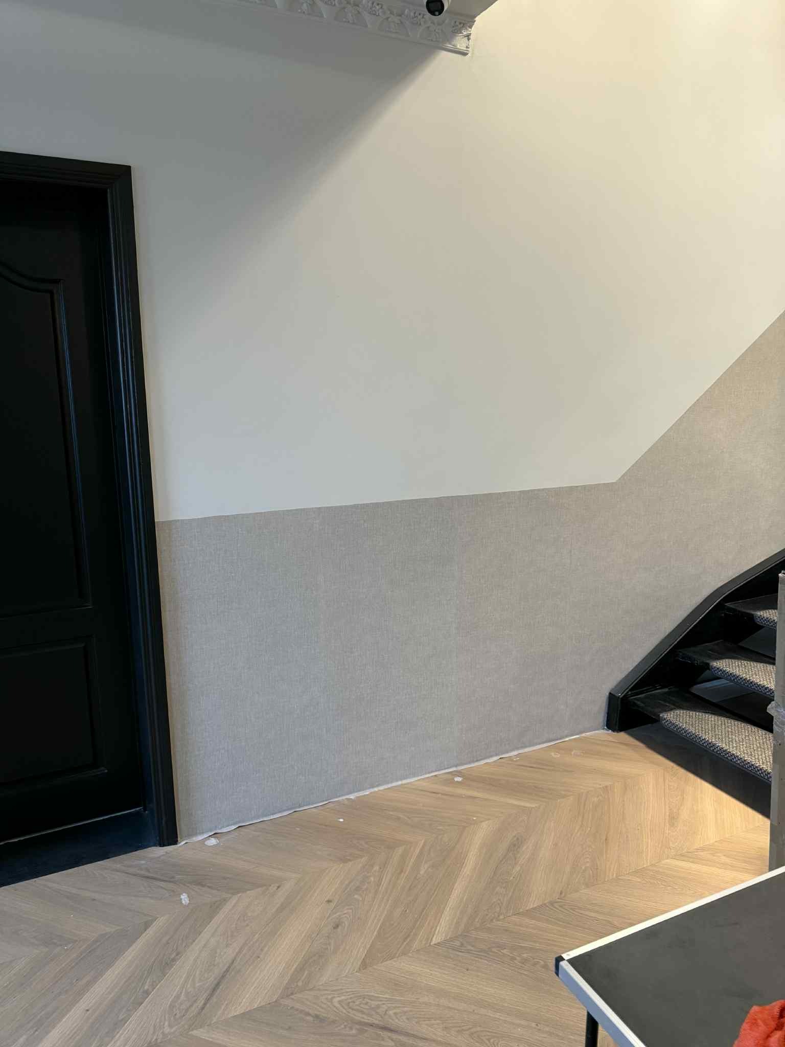 Wykończone wnętrze klatki schodowej: szara tapeta do połowy ściany, parkiet w jodełkę, czarne schody i drzwi. Widoczne detale wykończeniowe.