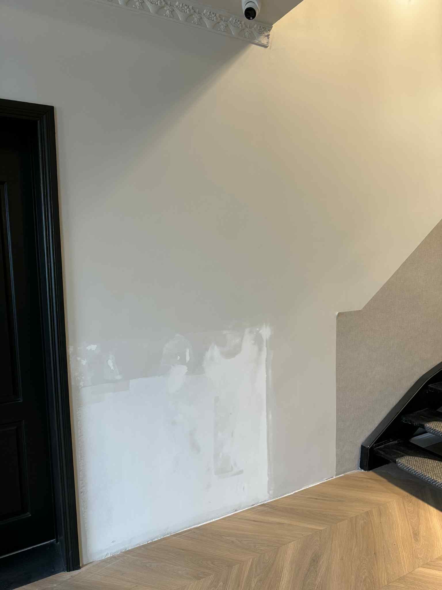 Ściana w trakcie przygotowania do malowania, widoczne ślady szpachlowania. Parkiet ułożony w jodełkę, czarne drzwi po lewej stronie kadru, fragment schodów po prawej.