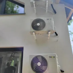 Trzy klimatyzatory marki Noa zamontowane jeden nad drugim na zewnętrznej ścianie budynku, widoczne białe wsporniki i czarne peszle, okna z refleksami wnętrza w tle.