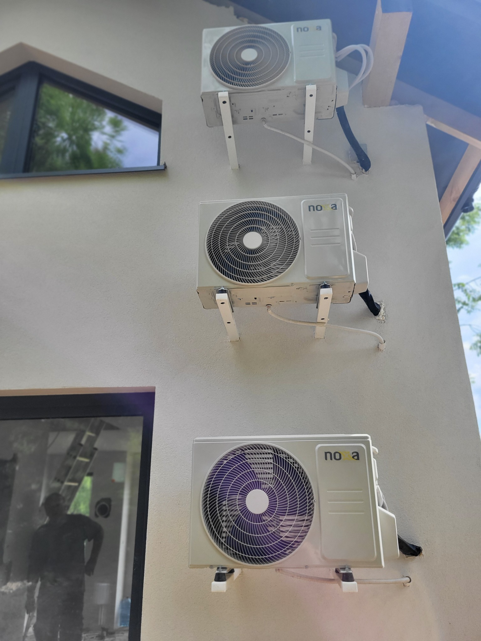 Trzy klimatyzatory marki Noa zamontowane jeden nad drugim na zewnętrznej ścianie budynku, widoczne białe wsporniki i czarne peszle, okna z refleksami wnętrza w tle.