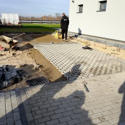 Mariusz Jach JACHKOP Kompleksowe usługi minikoparką - Układanie ażurowych płyt betonowych na podsypce piaskowej. W tle pracownik zagęszcza podłoże ubijarką. Widoczne narzędzia i materiały budowlane.