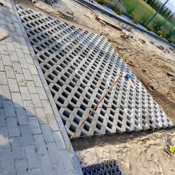 Mariusz Jach JACHKOP Kompleksowe usługi minikoparką - Układanie ażurowych płyt betonowych na piasku, widoczne narzędzia i fragment kostki brukowej. Prace w toku, teren przygotowany pod dalsze prace.