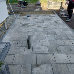 Mariusz Jach JACHKOP Kompleksowe usługi minikoparką - Układanie szarej kostki brukowej na posesji, widoczne narzędzia i częściowo ułożona nawierzchnia. Prace brukarskie w toku, zarys ogrodu w tle.