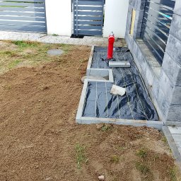 Mariusz Jach JACHKOP Kompleksowe usługi minikoparką - Przygotowany teren pod trawnik z rolki, widoczna czarna mata, obrzeża z kostki, hydrant i brama w tle. Częściowo wyrównana ziemia.