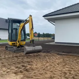 Ż&oacute;łta mini koparka na przygotowanym terenie budowy, obok nowoczesny dom z szarą dach&oacute;wką i białymi ścianami, w tle widoczny kolejny dom.