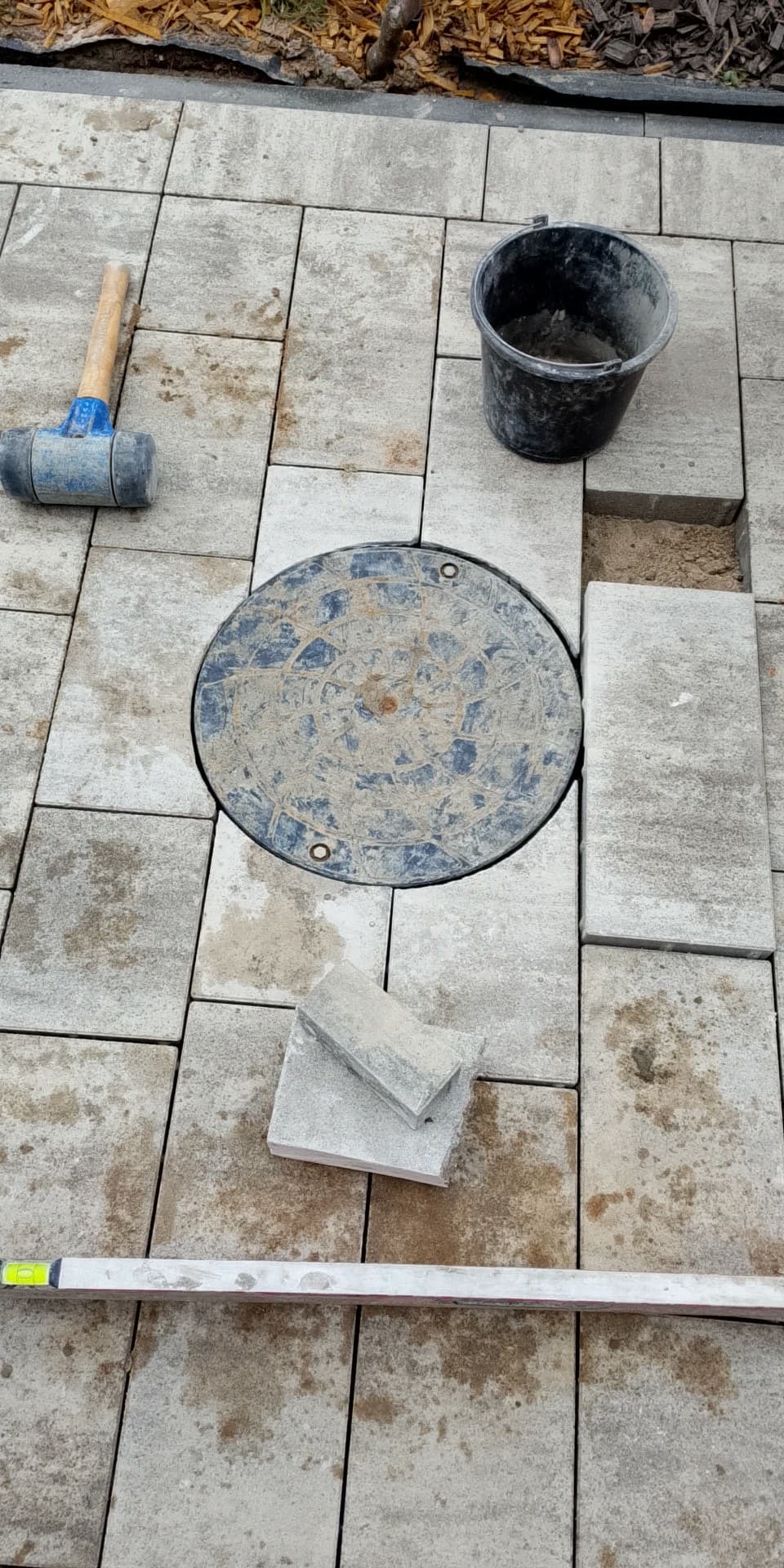 Układanie jasnoszarej kostki brukowej wokół okrągłej studzienki. Widoczny poziomica, gumowy młotek i wiadro. Częściowo ułożona nawierzchnia.