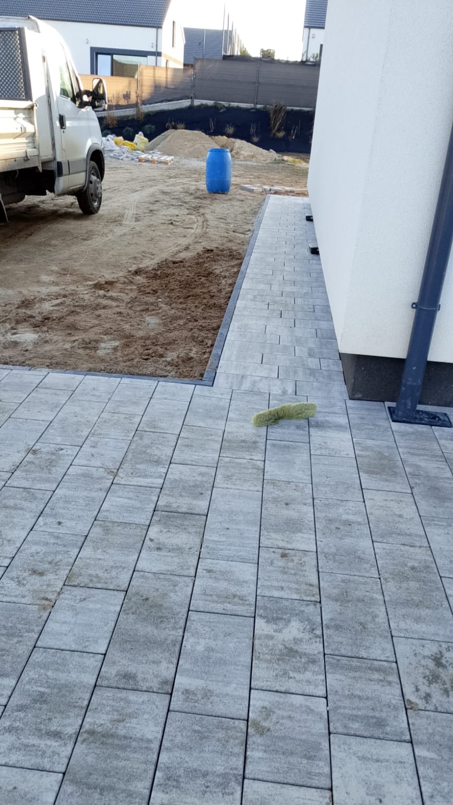 Ułożona kostka brukowa w odcieniach szarości, z widocznym fragmentem posesji w tle i narzędziem do czyszczenia na pierwszym planie. Widoczny fragment samochodu dostawczego.