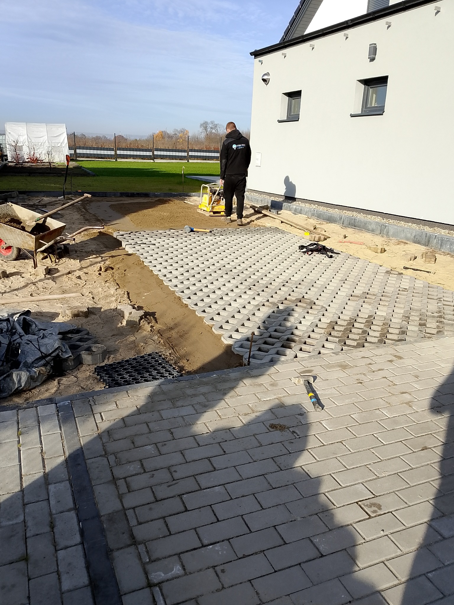 Układanie ażurowych płyt betonowych na podsypce piaskowej. W tle pracownik zagęszcza podłoże ubijarką. Widoczne narzędzia i materiały budowlane.