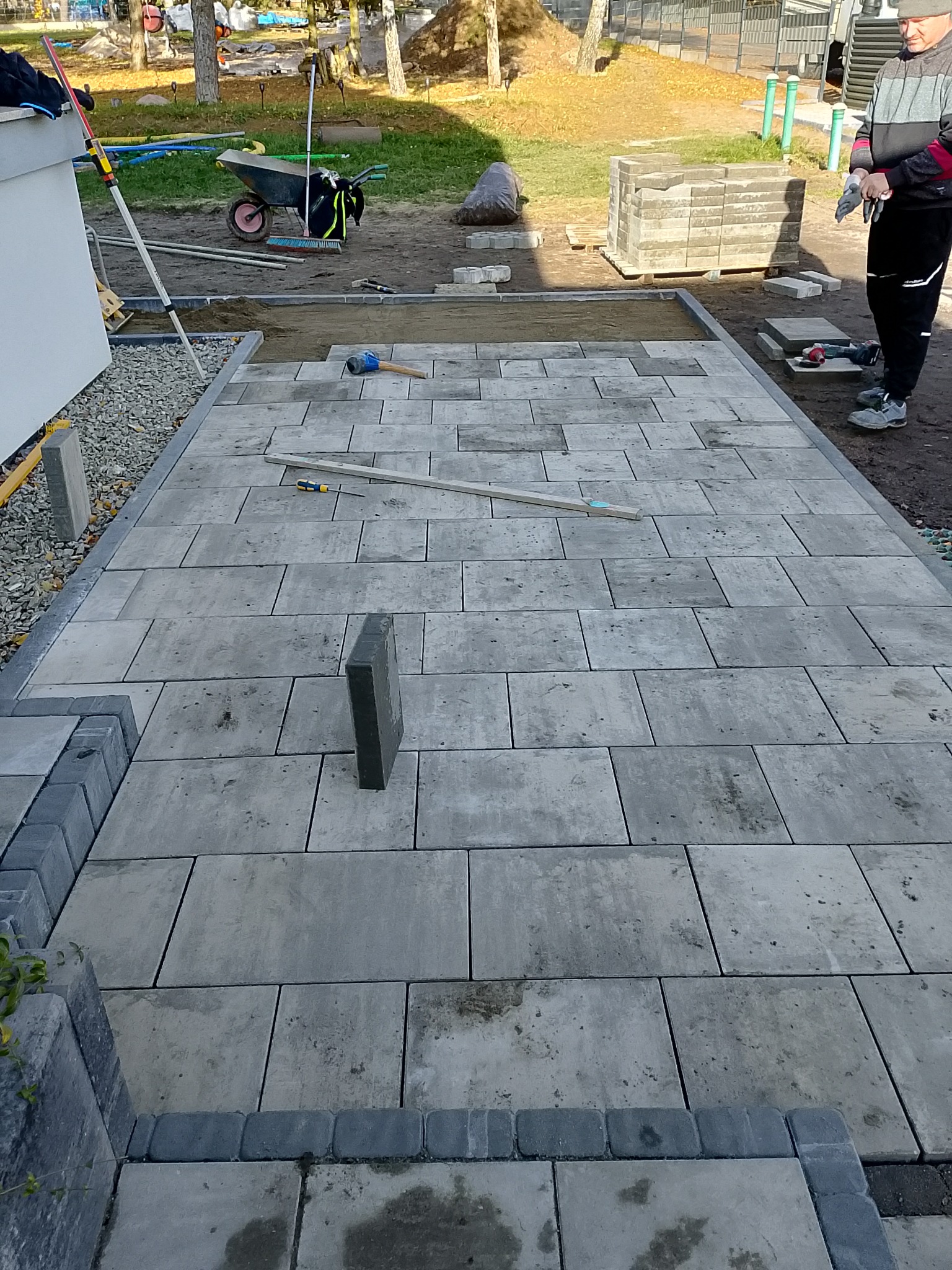 Układanie szarej kostki brukowej na posesji, widoczne narzędzia i częściowo ułożona nawierzchnia. Prace brukarskie w toku, zarys ogrodu w tle.