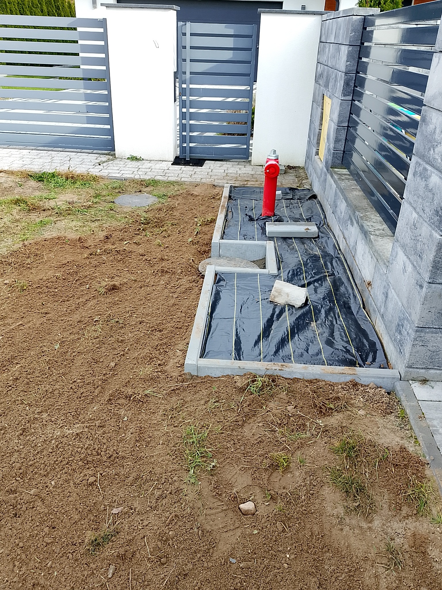 Przygotowany teren pod trawnik z rolki, widoczna czarna mata, obrzeża z kostki, hydrant i brama w tle. Częściowo wyrównana ziemia.