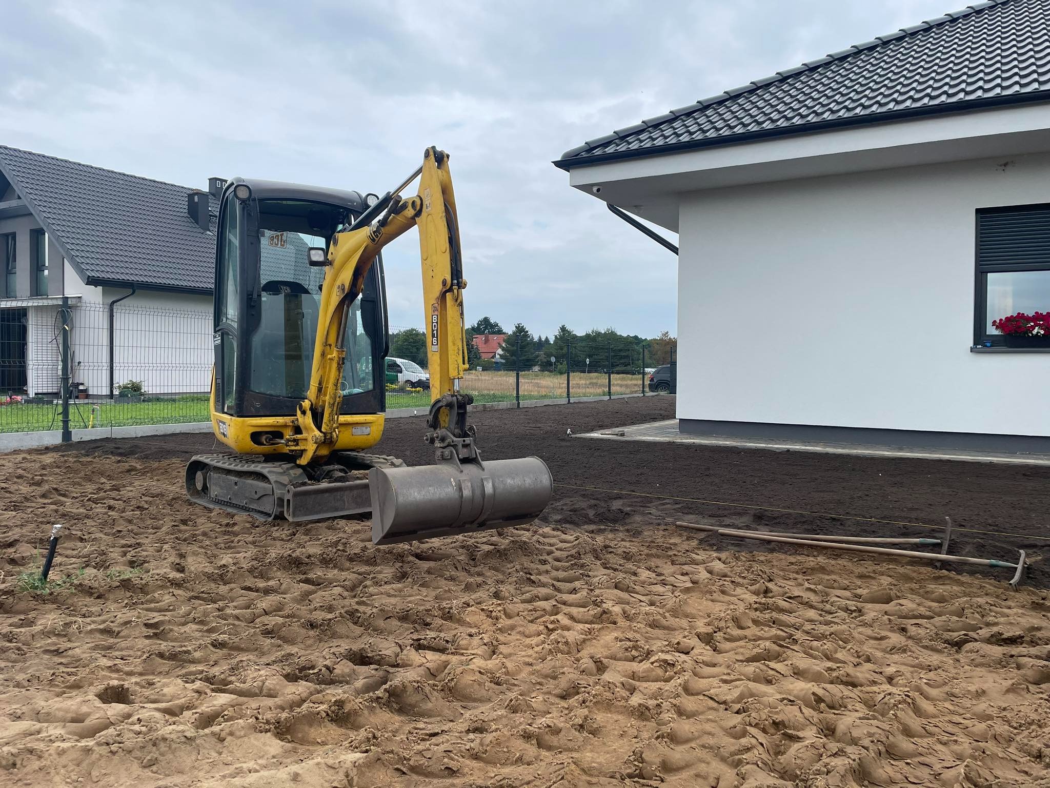 Żółta mini koparka na przygotowanym terenie budowy, obok nowoczesny dom z szarą dachówką i białymi ścianami, w tle widoczny kolejny dom.