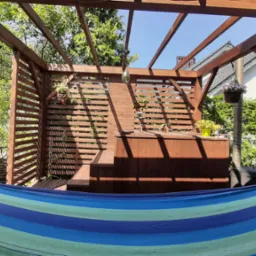 Drewniana konstrukcja altany z elementami ażurowymi, widok z wnętrza, hamak w pierwszym planie, rośliny w tle.