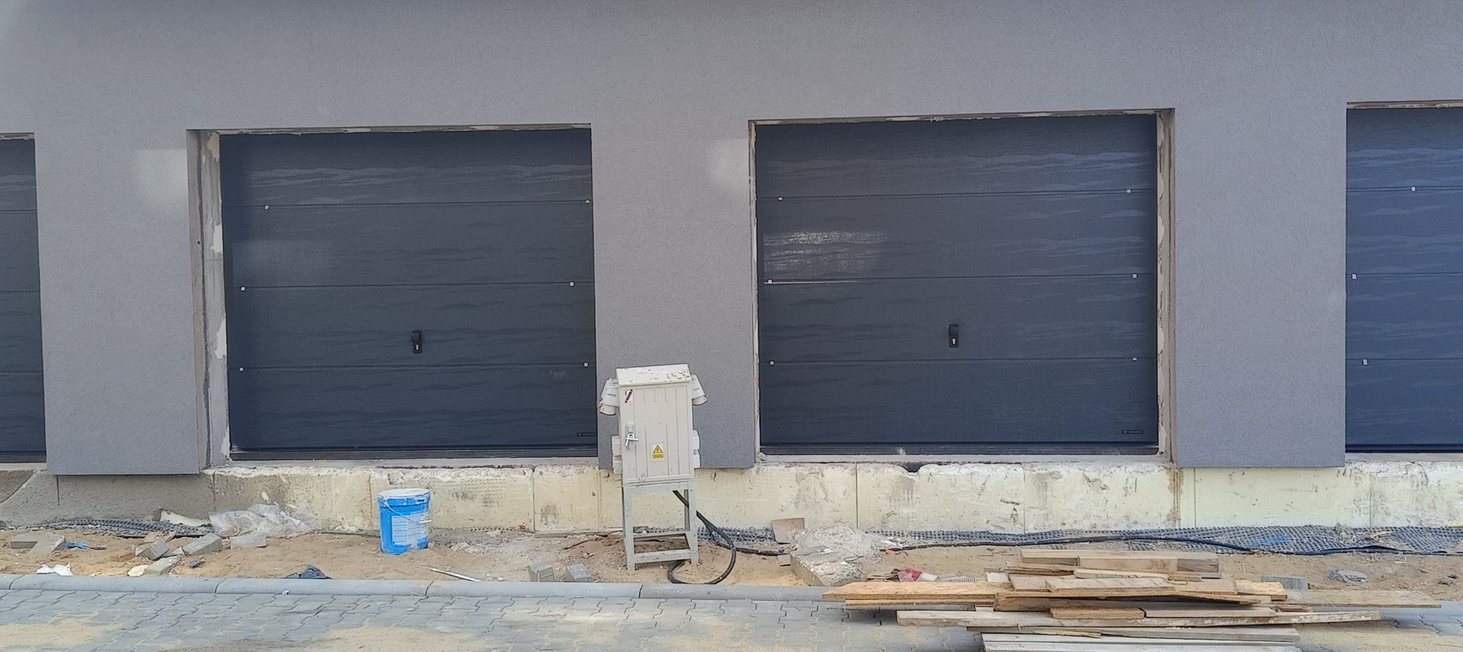 Montaż bram garażowych segmentowych UniPro w kolorze antracyt na nowej deweloperce, widoczna skrzynka elektryczna i pozostałości po pracach budowlanych.