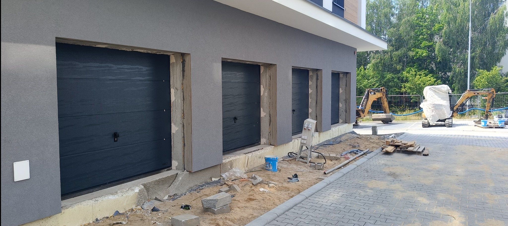 Montaż trzech segmentowych bram garażowych w szarym budynku, widoczne przygotowane otwory i teren budowy z maszynami w tle.