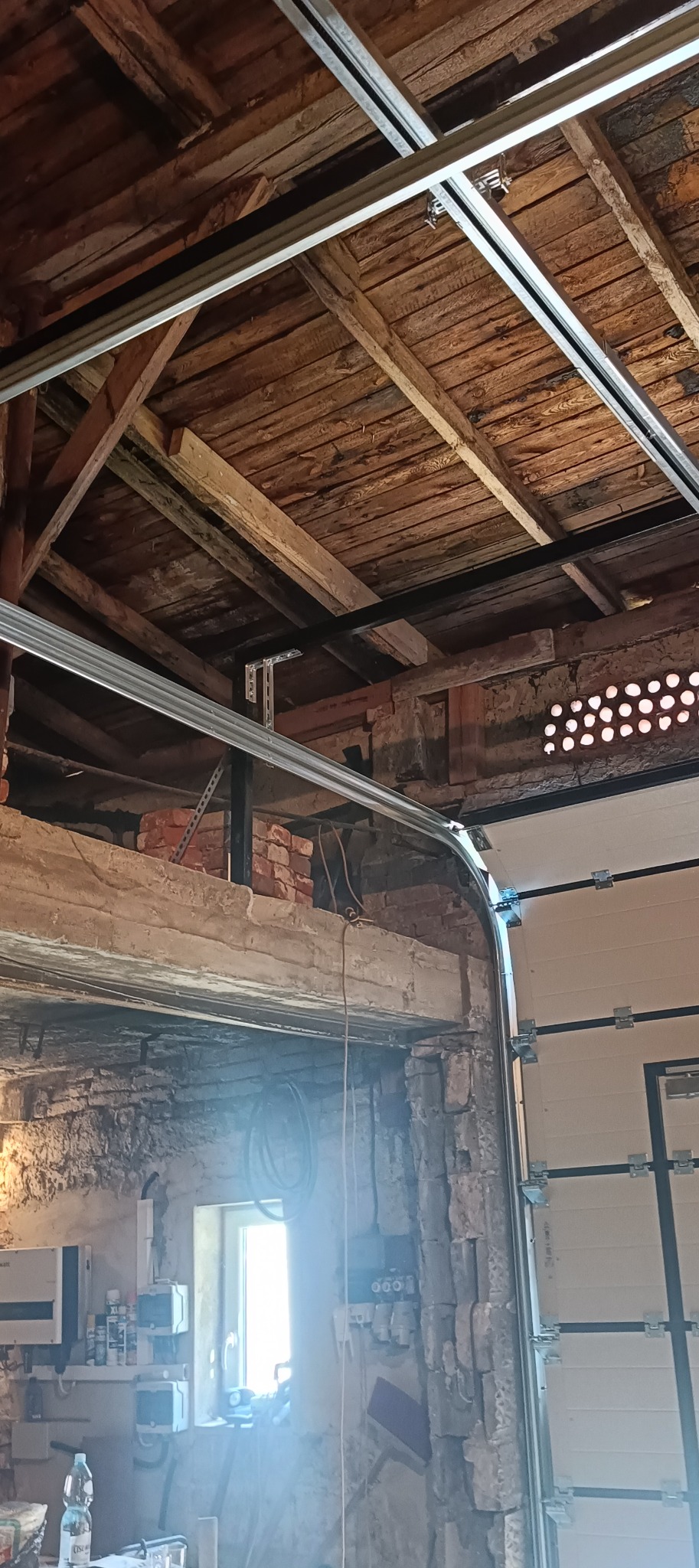 Montaż bramy garażowej z widoczną konstrukcją stalową podtrzymującą szyny, w garażu z surowym, ceglanym wykończeniem ścian i drewnianym stropem.