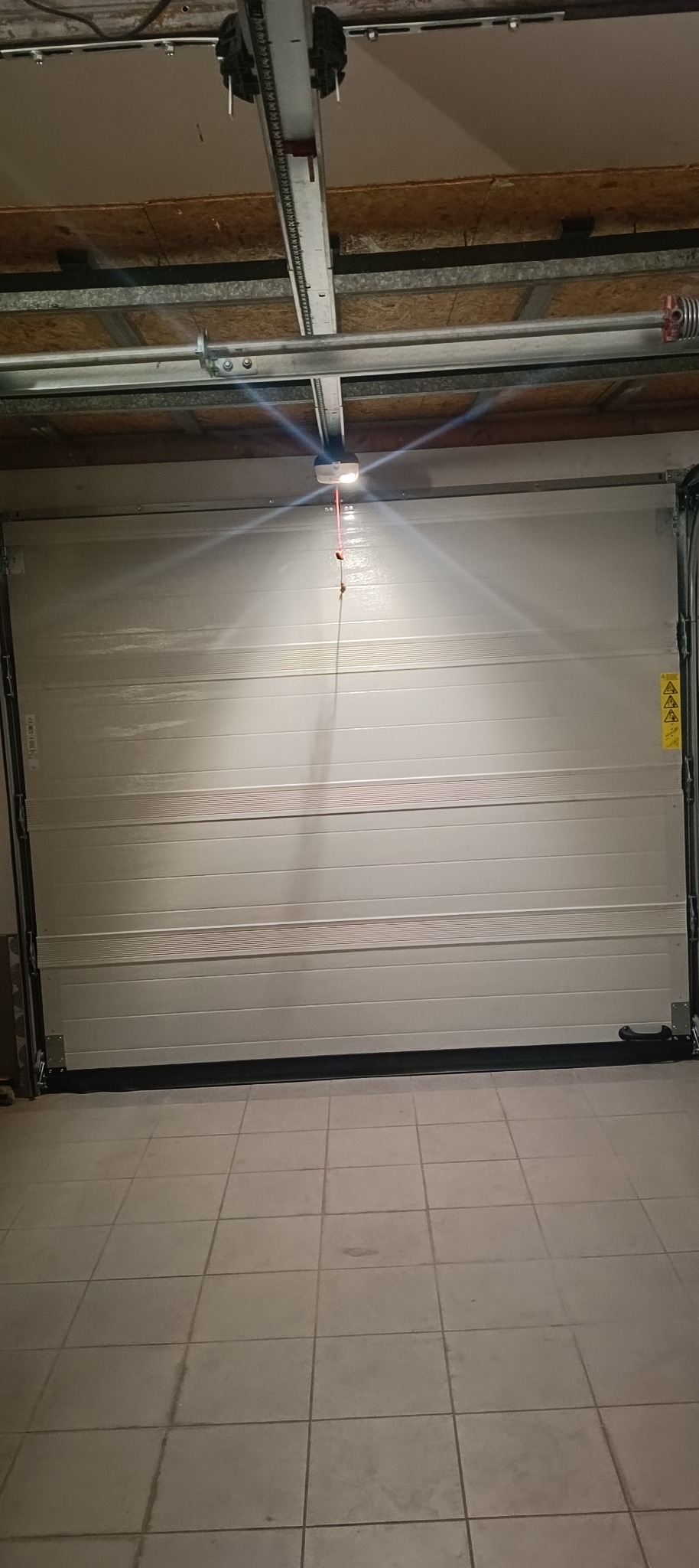 Zamknięta biała segmentowa brama garażowa z widocznym mechanizmem napędowym i oświetleniem wewnątrz garażu, widok z perspektywy podłogi z płytek.