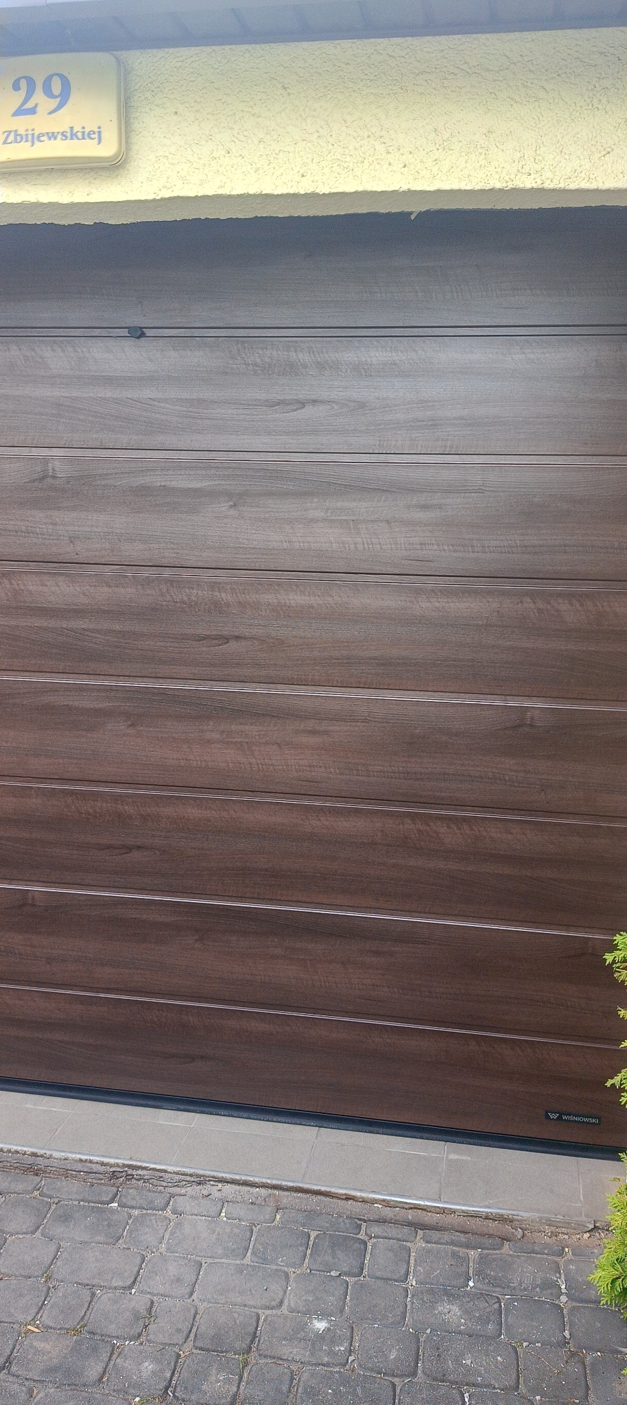 Zamknięta, segmentowa brama garażowa w odcieniach brązu z widocznym adresem budynku na żółtej tabliczce powyżej.