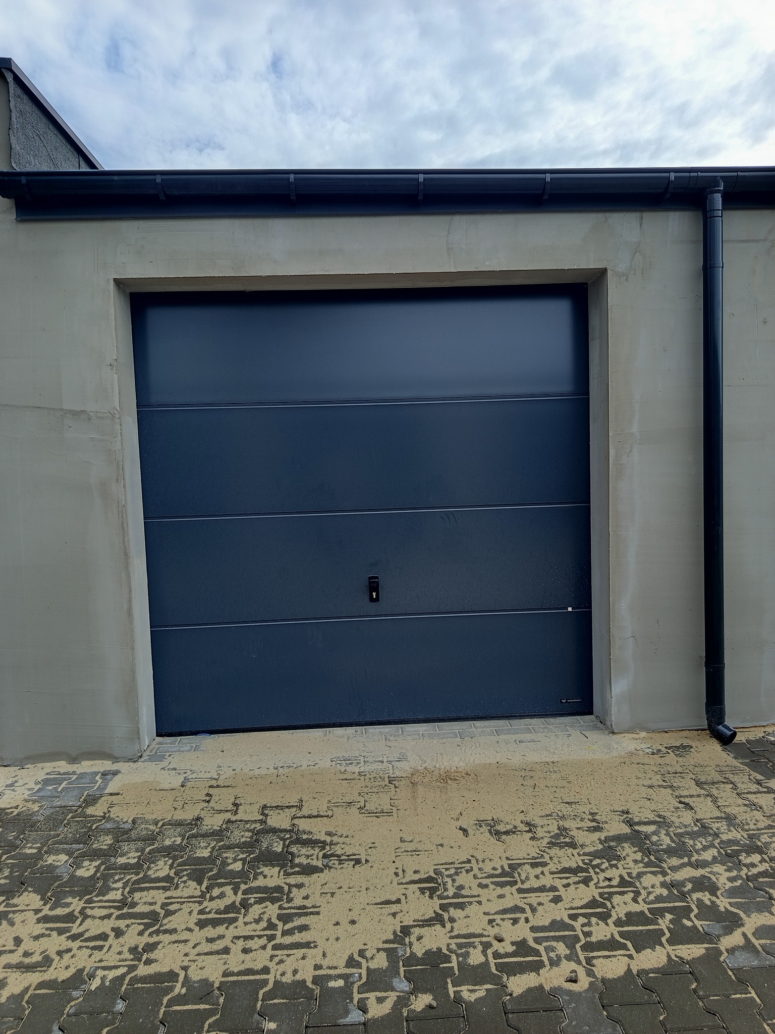 Ciemna brama garażowa segmentowa, z poziomymi przetłoczeniami, zamontowana w surowym, betonowym garażu. Na dole mokra kostka brukowa.