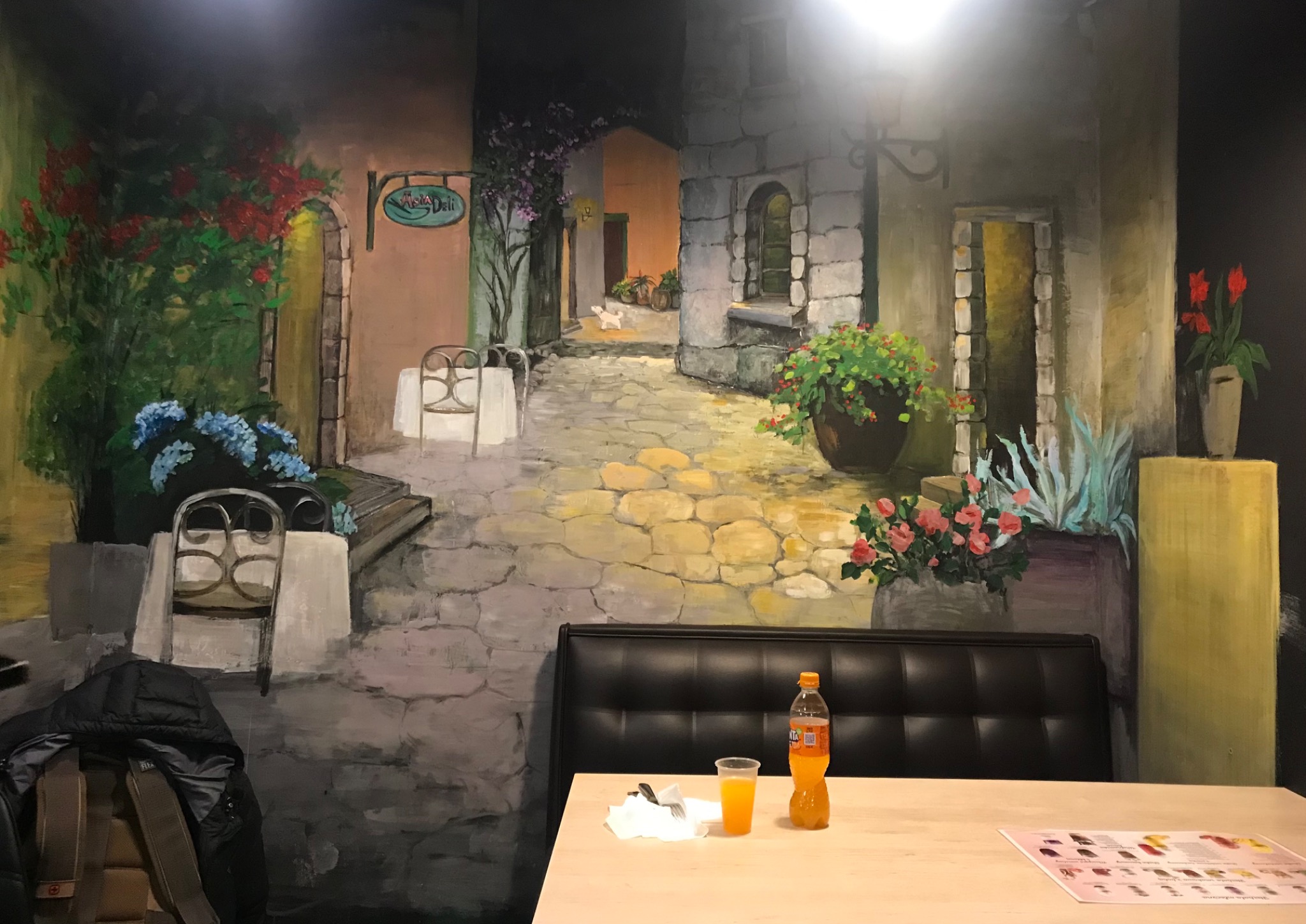 Ściana z ręcznie malowanym pejzażem miejskim w stylu włoskim, z widocznym stolikiem i krzesłami, tworząca iluzję głębi w restauracji, z fragmentem stolika i napojem w pierwszym planie.