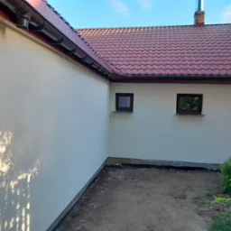 Świeżo otynkowana ściana zewnętrzna domu z widocznymi oknami, rynnami i fragmentem dachu z dachówki ceramicznej w odcieniach czerwieni, z lekkim cieniowaniem od liści drzewa.