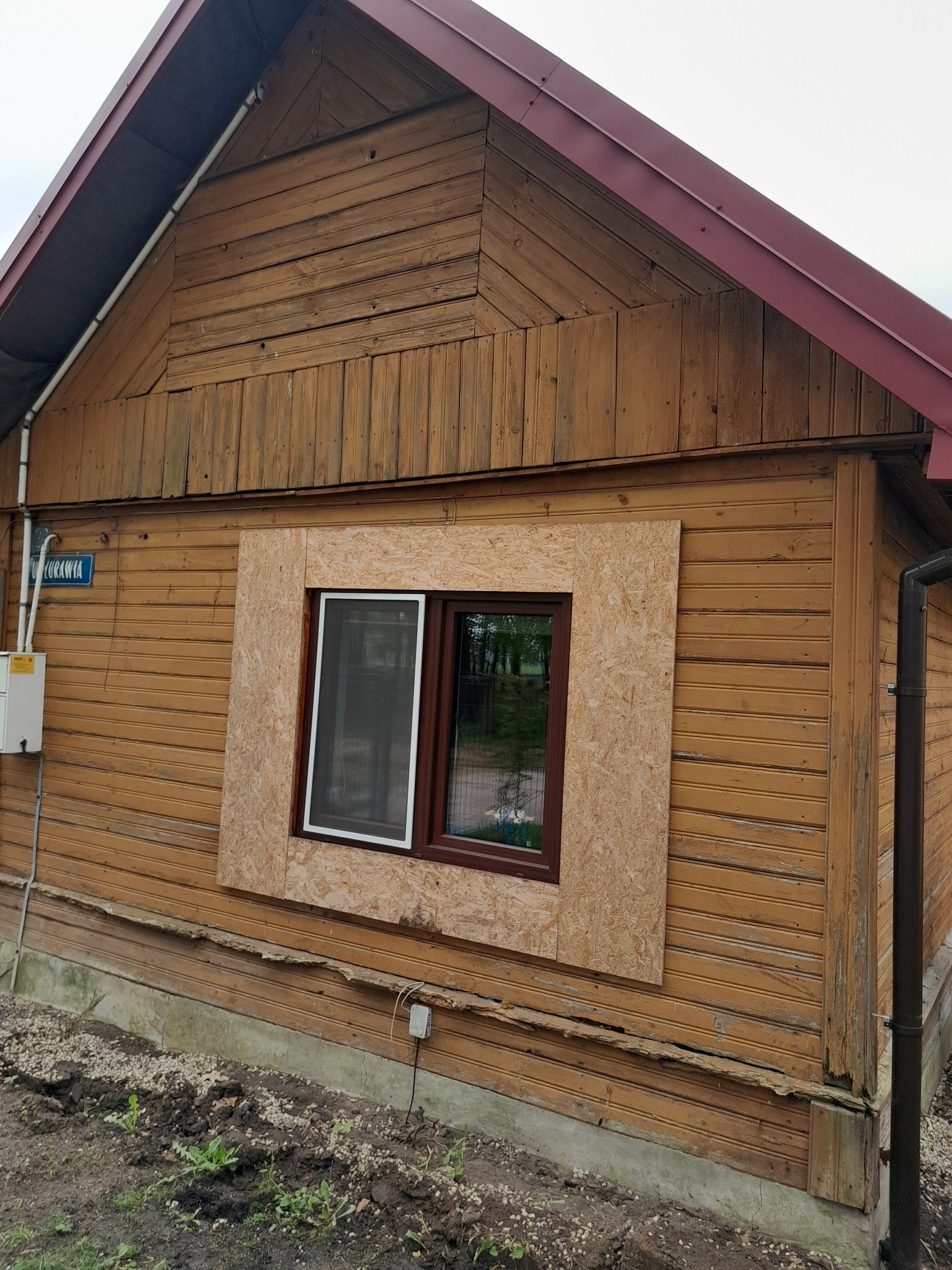 Fragment drewnianej elewacji domu w trakcie remontu, z oknem obramowanym płytą OSB przed dalszymi pracami wykończeniowymi.