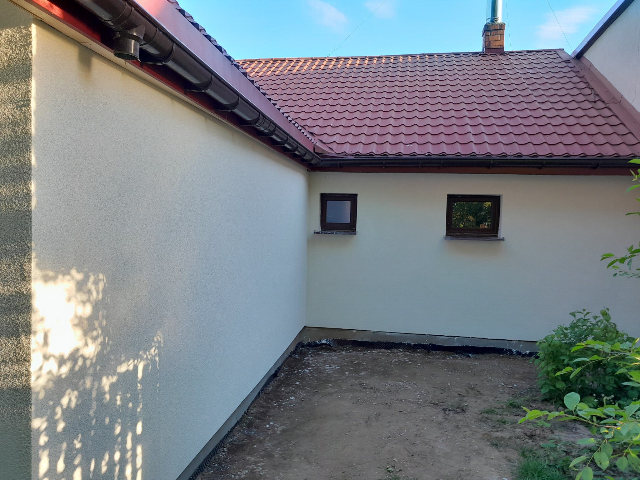 Świeżo otynkowana ściana zewnętrzna domu z widocznymi oknami, rynnami i fragmentem dachu z dachówki ceramicznej w odcieniach czerwieni, z lekkim cieniowaniem od liści drzewa.