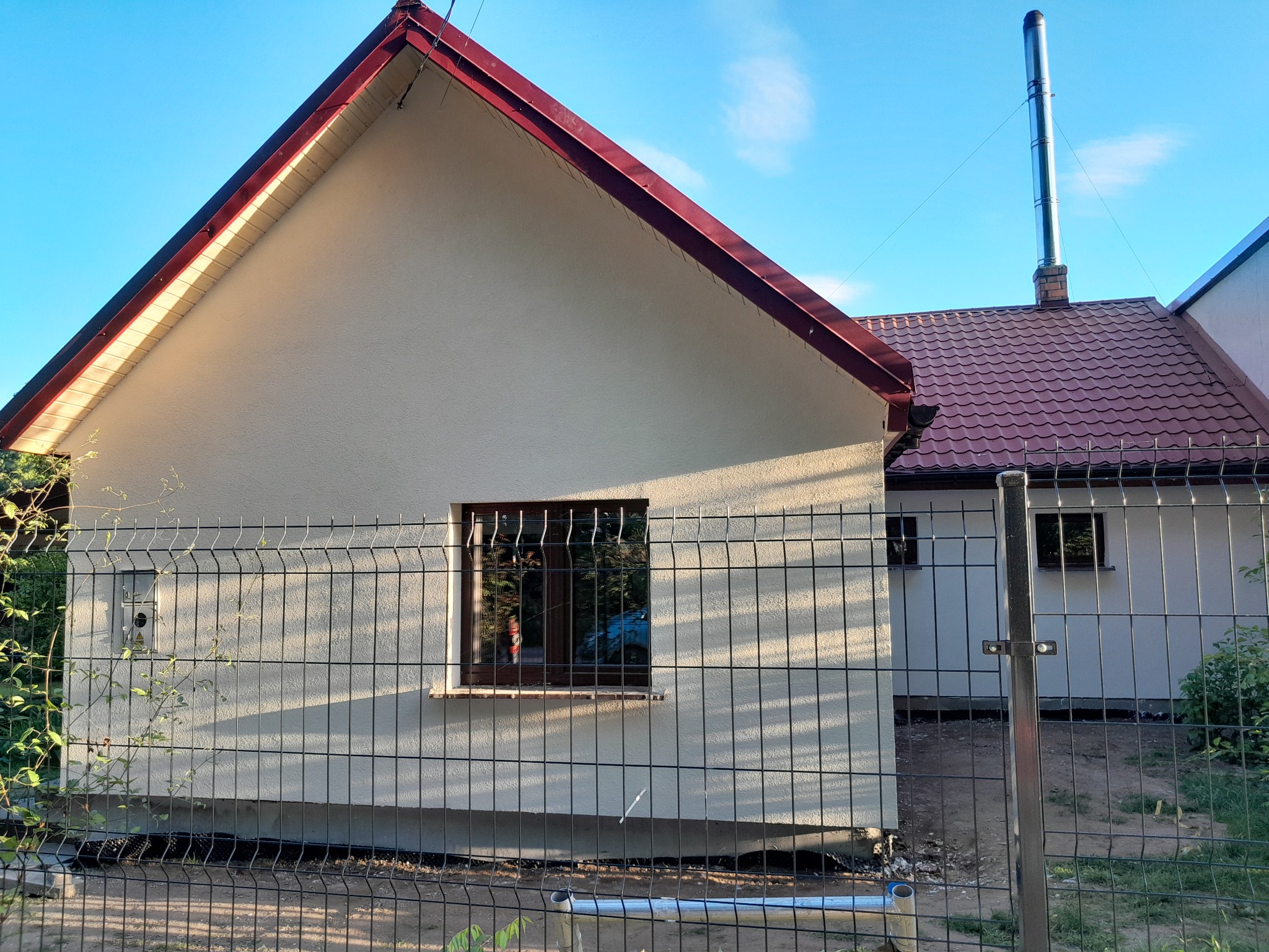 Świeżo wykończona elewacja domu jednorodzinnego w jasnym kolorze, widoczna przez metalową siatkę ogrodzeniową, z brązowymi ramami okien i czerwonym dachem, wysoki komin ze stali nierdzewnej.