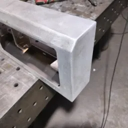 Szlifowana, aluminiowa rama konstrukcyjna z wewnętrznymi wspornikami, leżąca na perforowanym stole warsztatowym, widoczny przewód zasilający i narzędzia w tle.